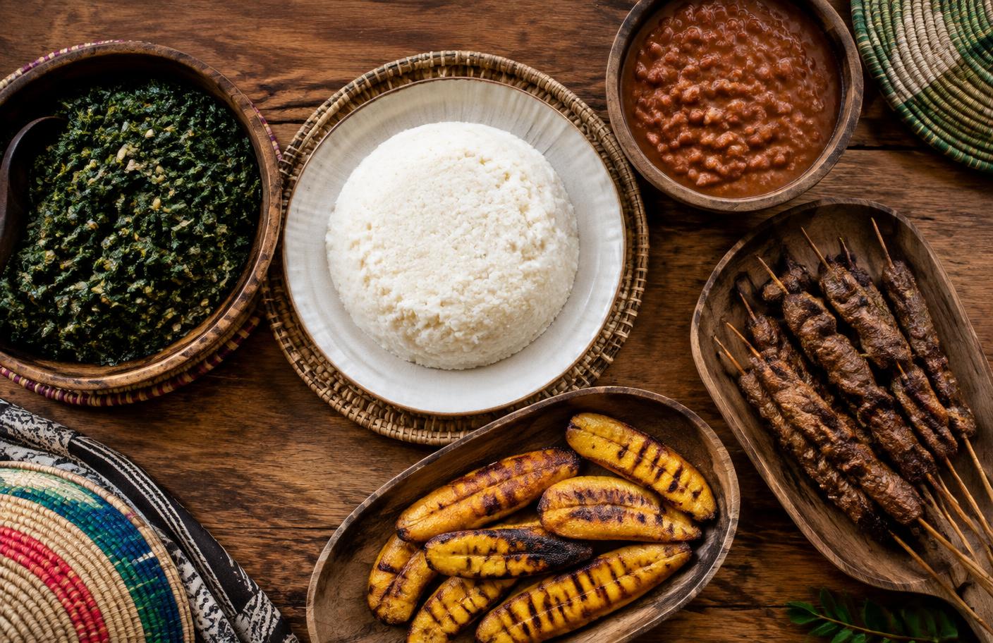 Isombe, ugali, beans, grilled plantain and brochettes on a wooden table
